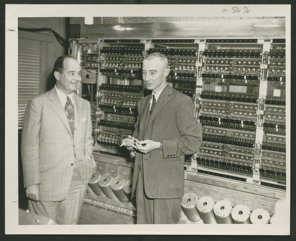 Two men in suits standing in front of an early computer. The computer takes up the entire wall behind them and consists of a series of large tubes at the bottom and exposed circuits above.